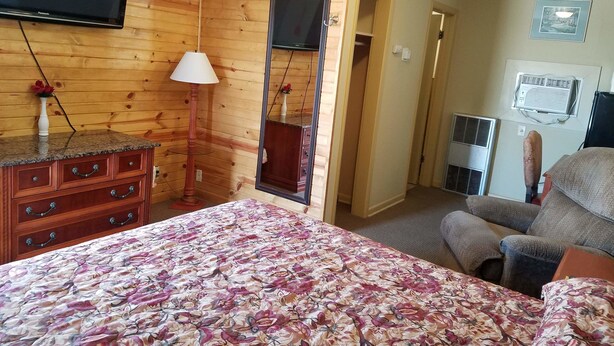 Une pièce aux murs en bois, avec un lit recouvert d’une couvre-lit à motifs floraux, une commode, un téléviseur, une lampe et une chaise.