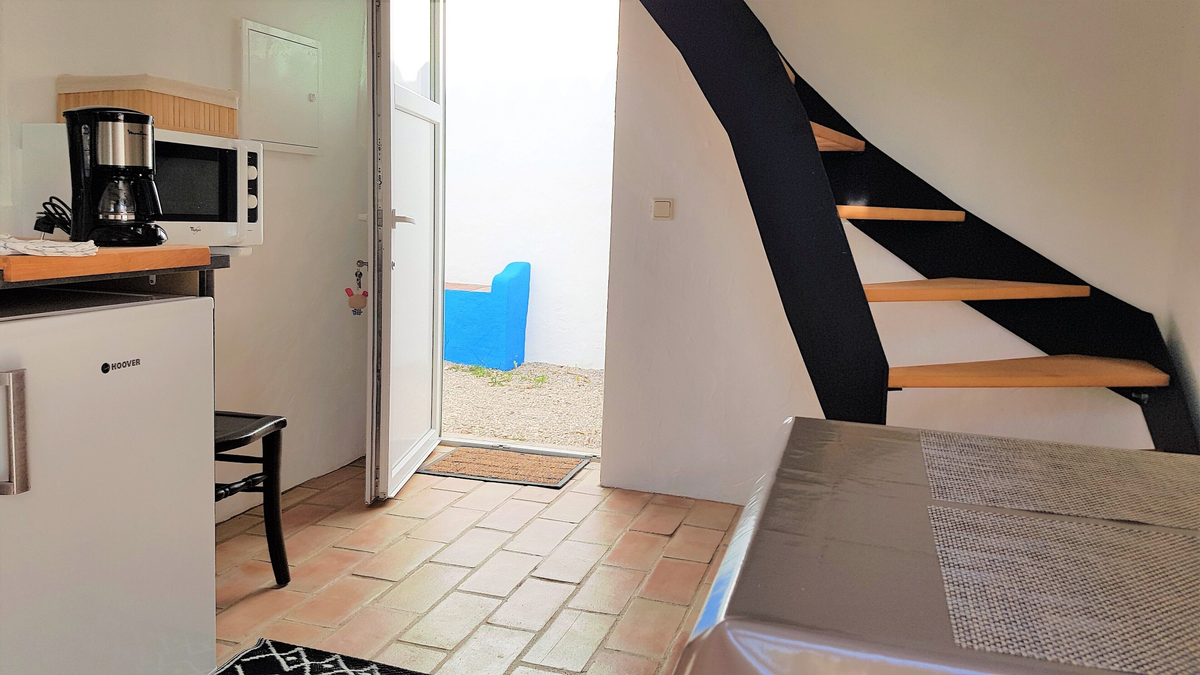 A compact kitchen with a microwave, coffee maker, and a refrigerator. A wooden staircase leads to a balcony with a blue chair.