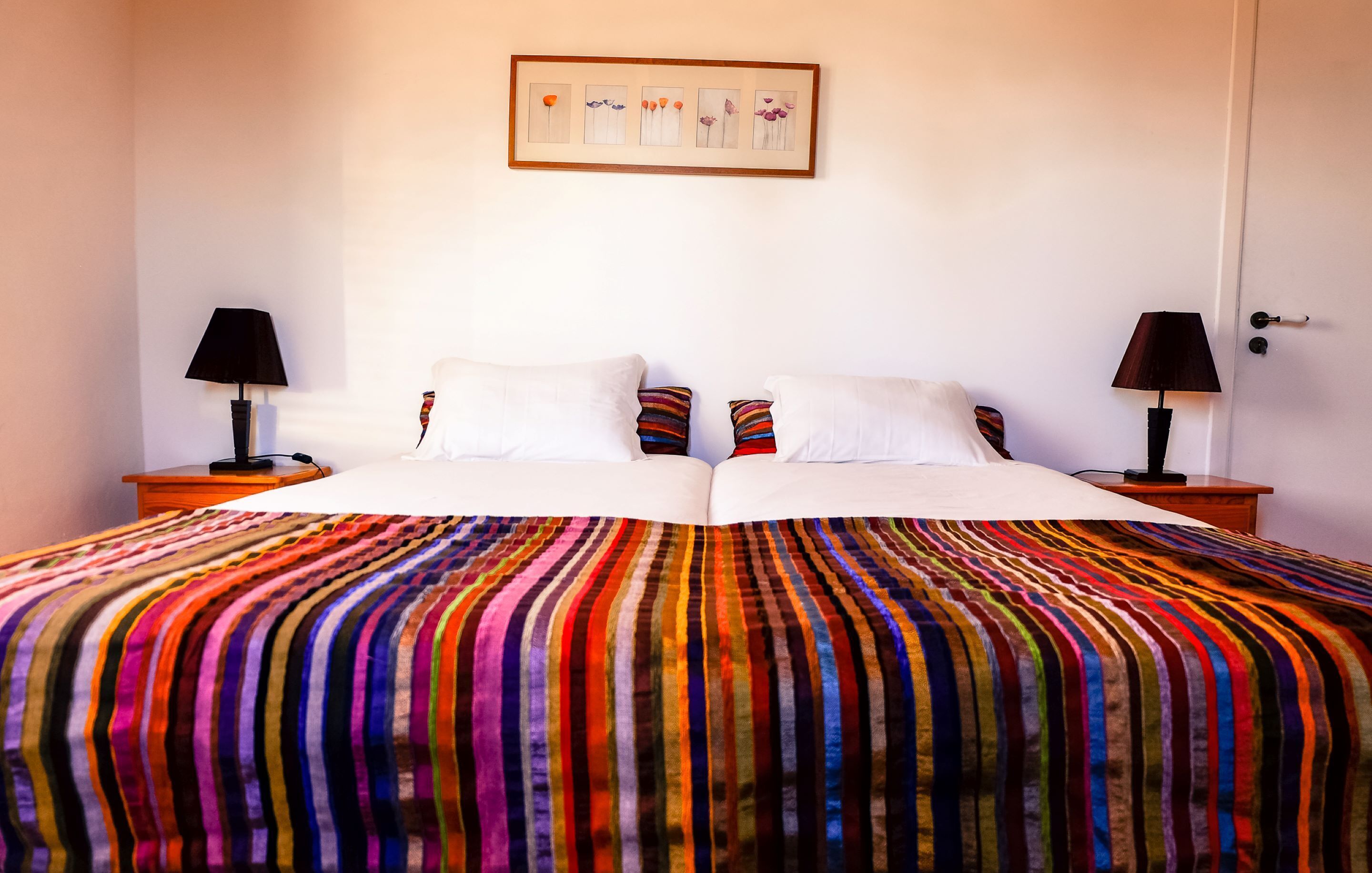 A double bed with a colorful striped bedspread, two bedside tables with lamps, and a framed artwork on the wall.
