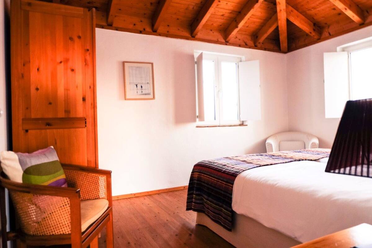 A bedroom with a wooden ceiling, a bed with a striped blanket, a wicker chair, and a white chair.