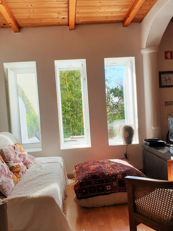 A bedroom with a bed, a chair, and a desk. There are three windows providing views of greenery and a building. - Quinta dos Cochichos – Country Houses (Olhao)