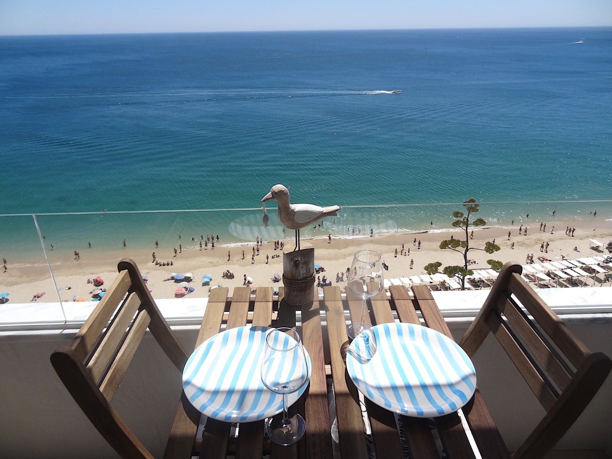 A balcony with wooden chairs and tables, overlooking a beach with people and a seagull.