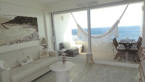 A modern living room with a white sofa, a round coffee table, and a dining set. A large window offers a view of the sea and a hammock. - Lingon Silver Apartment, Wifi, AC, Balcony (Sesimbra)