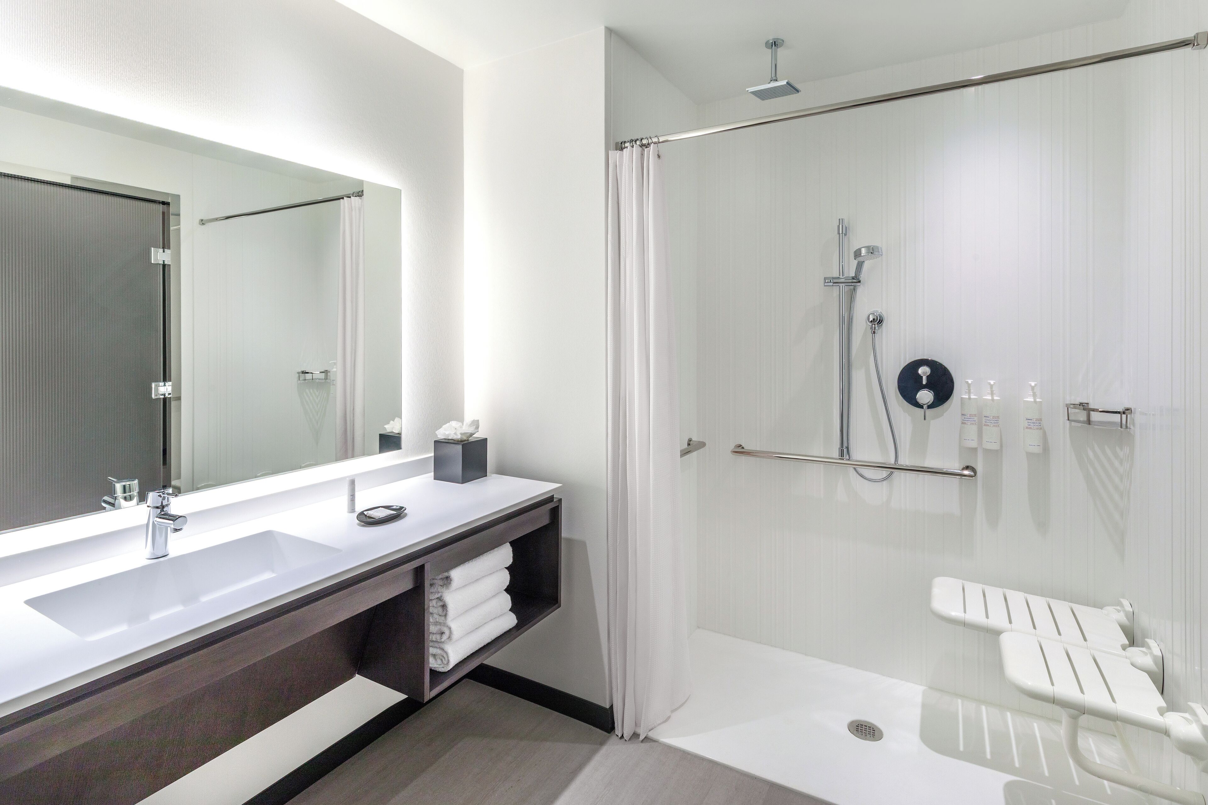 Un baño moderno con un espejo grande, un lavamanos, una ducha con puerta de cristal y un inodoro con asiento blanco.
