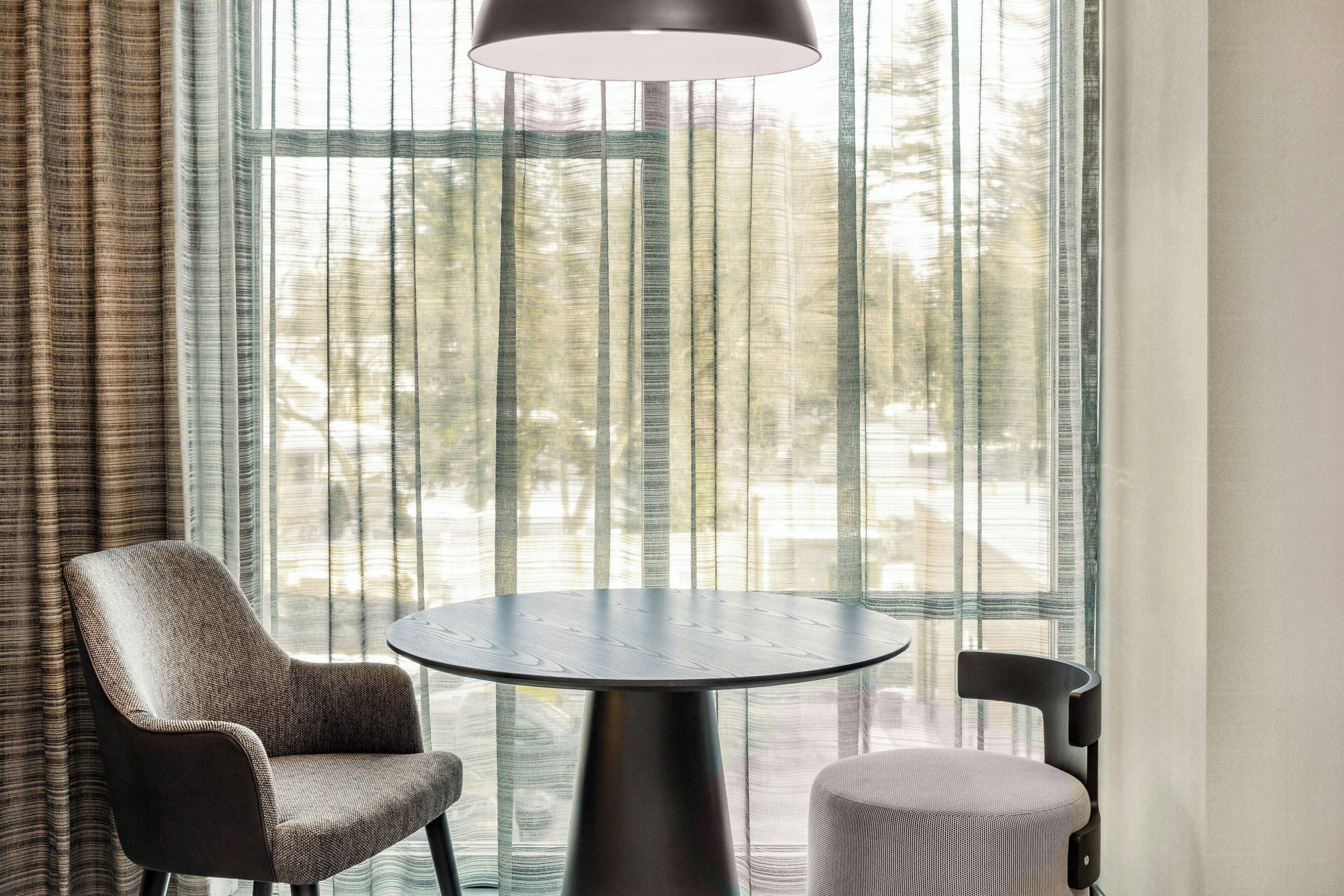 A room with a round table, two chairs, a lamp, and large windows with sheer curtains.