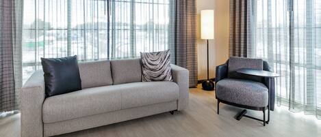 A modern living room with a sofa and armchair, large windows with sheer curtains, and a floor lamp.