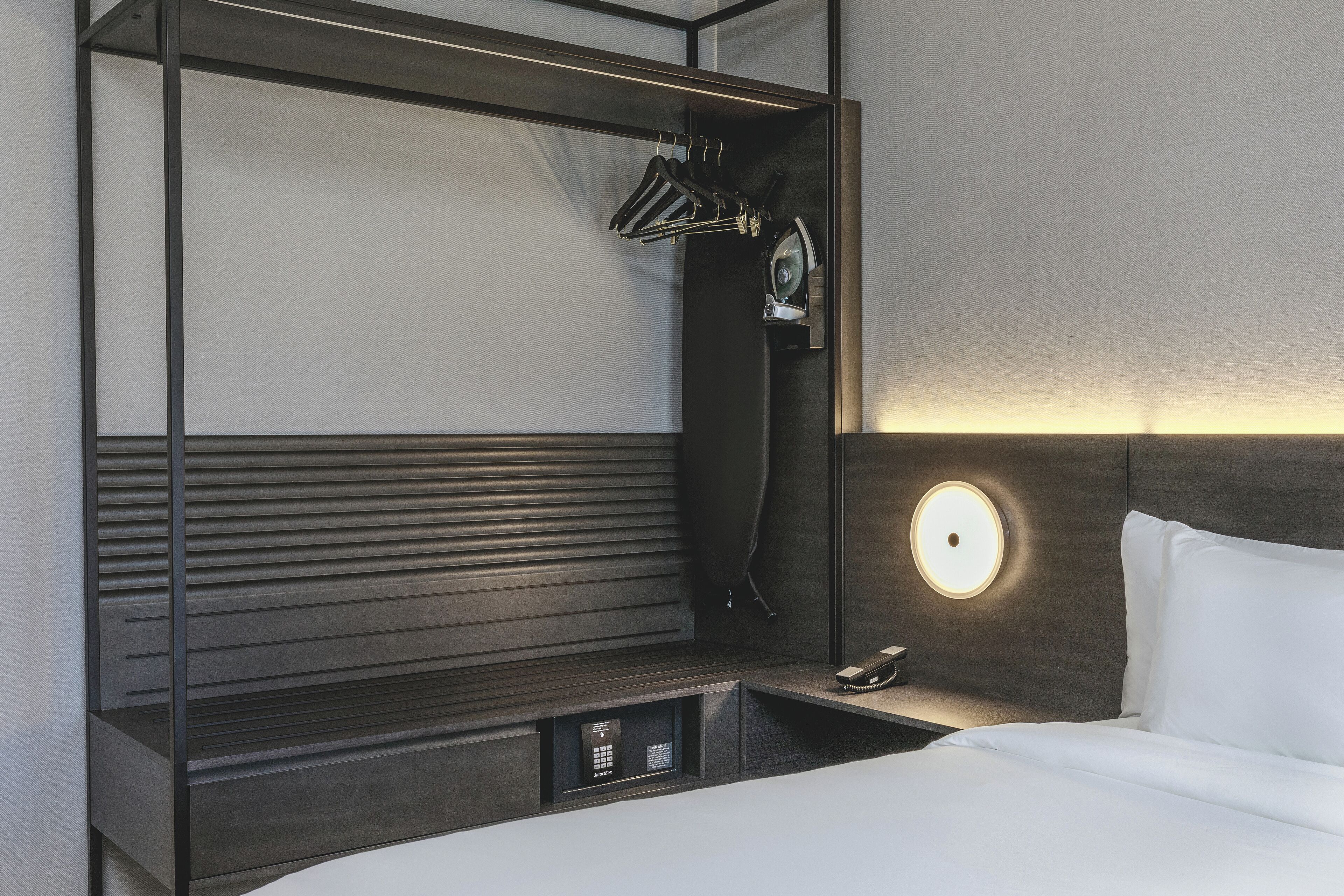 A modern hotel room with a bed, a bedside table with a lamp, a coat rack, and a built-in storage unit.