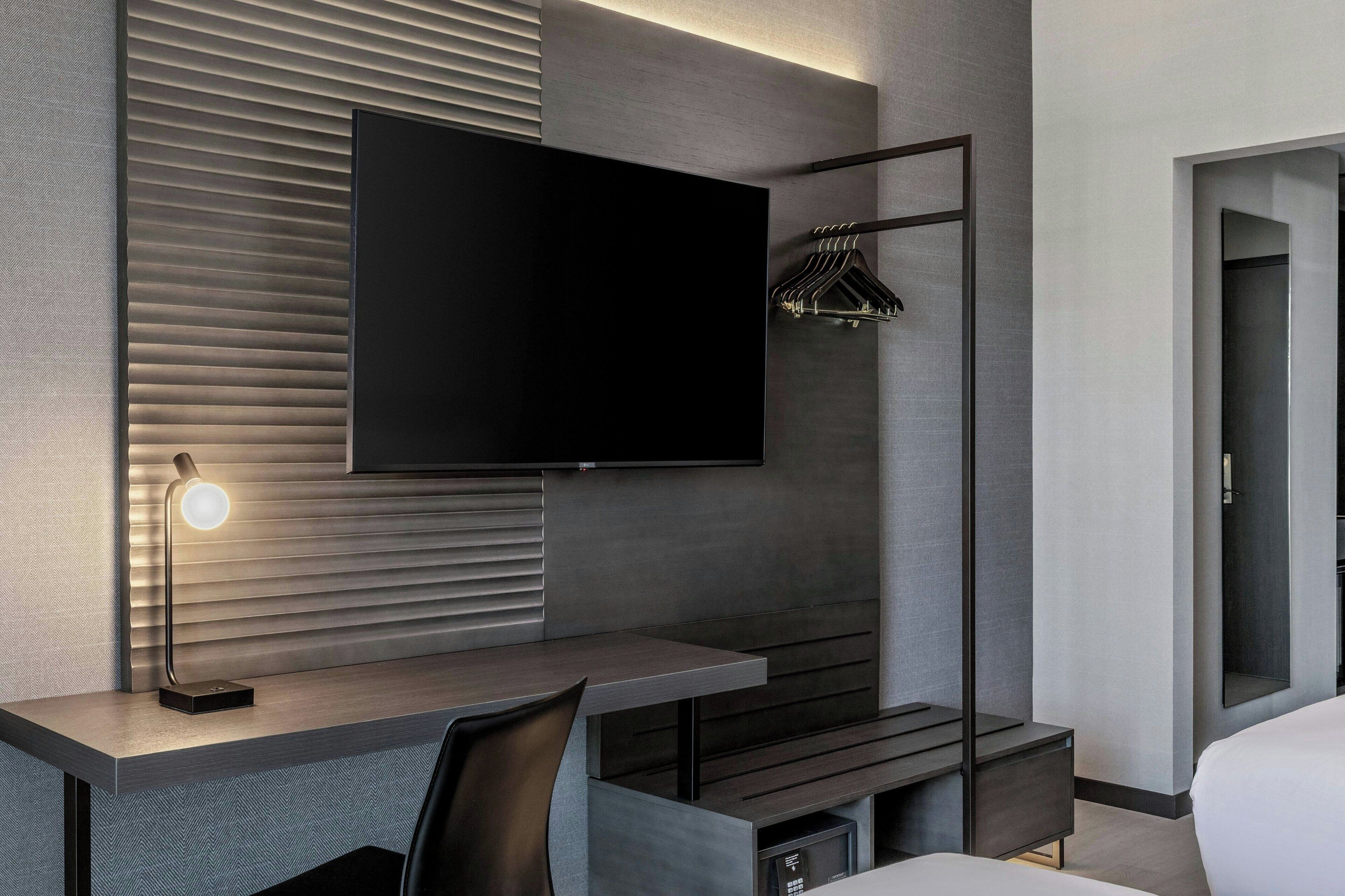 Una habitación de hotel moderna con una televisión de pantalla plana, un escritorio con lámpara, un perchero y una cama.
