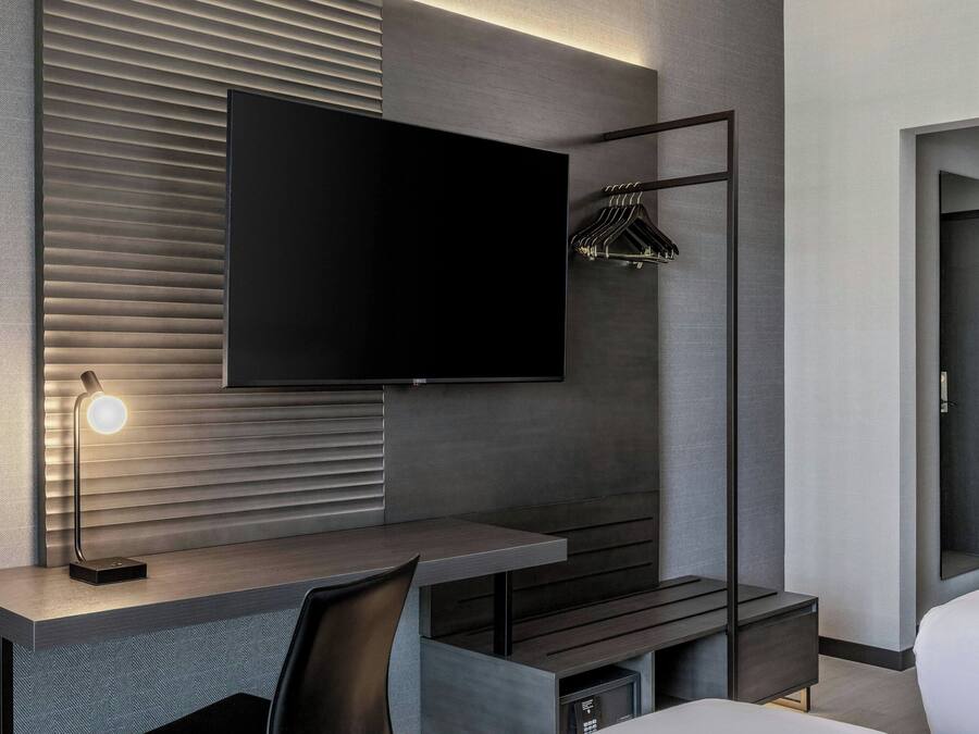 Una habitación de hotel moderna con una televisión de pantalla plana, un escritorio con lámpara, un perchero y una cama.