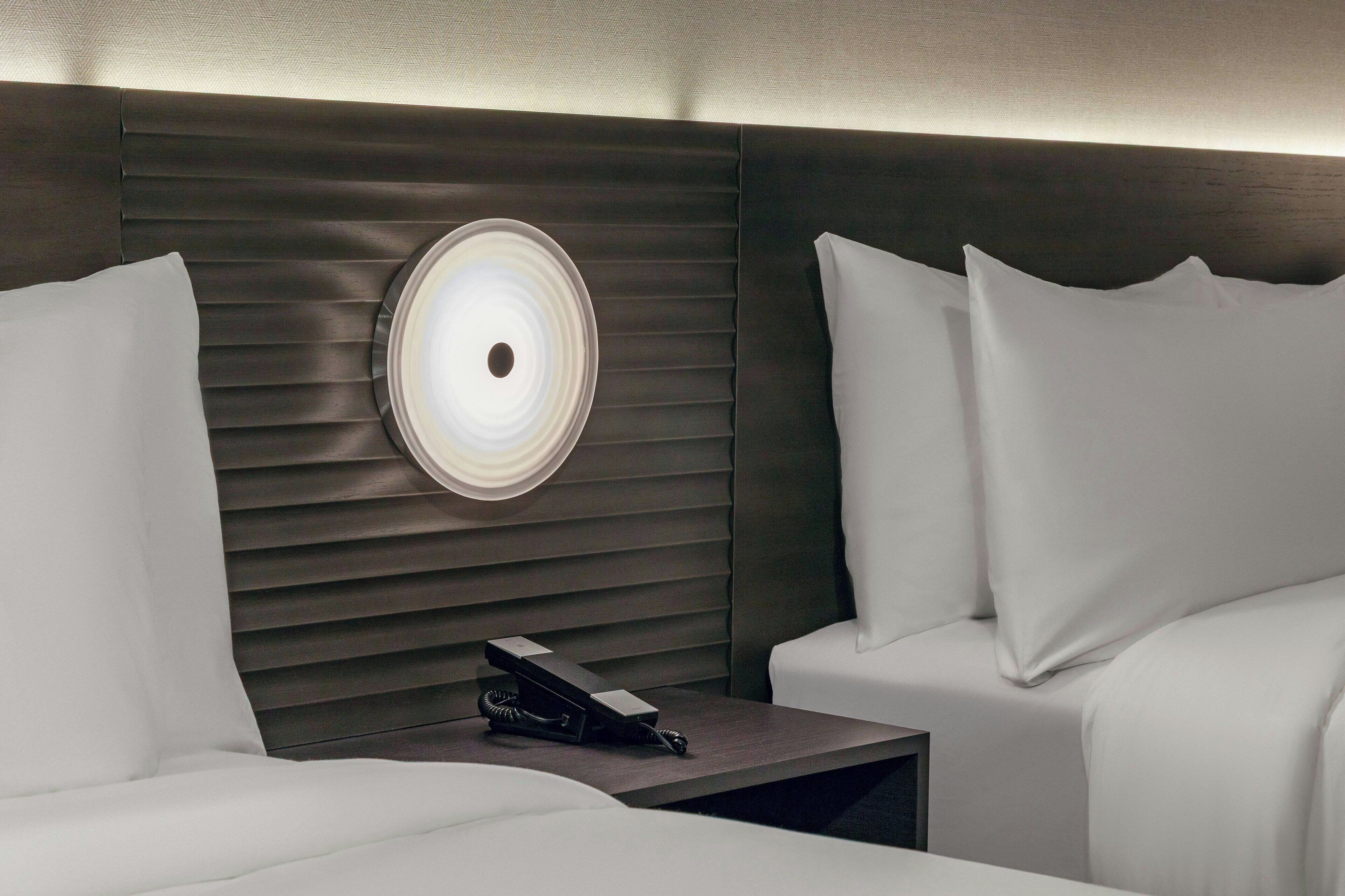 A hotel room with a modern design, featuring a bed with white bedding, a bedside table with a telephone, and a wall-mounted light fixture.