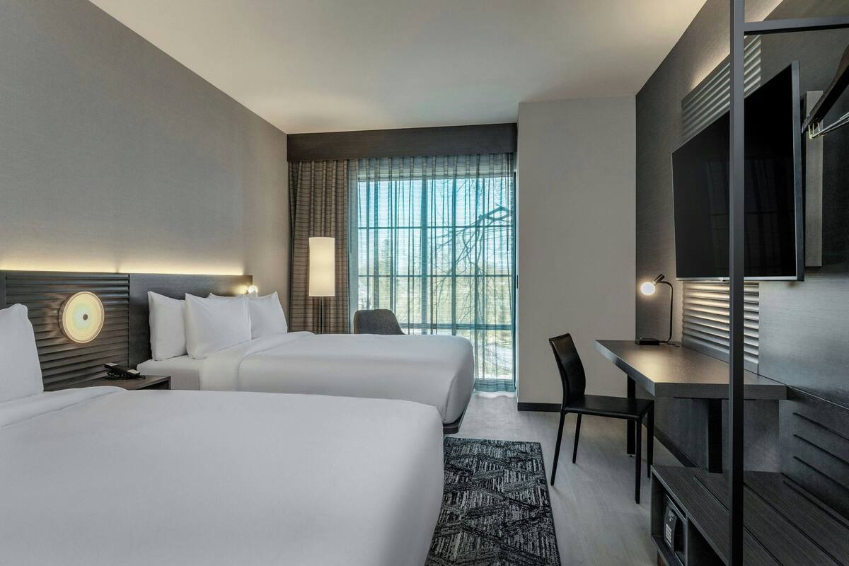 Habitación de hotel con dos camas, un escritorio, una silla, un televisor y un ventanal grande con persianas.