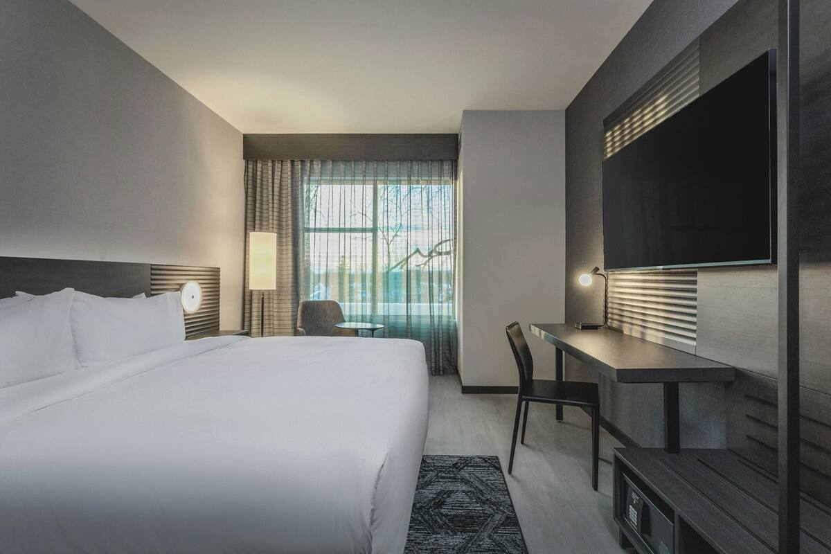Habitación de hotel con cama, escritorio, silla, televisor y ventana con cortinas.