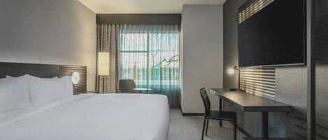 Habitación de hotel con cama, escritorio, silla, televisor y ventana con cortinas.