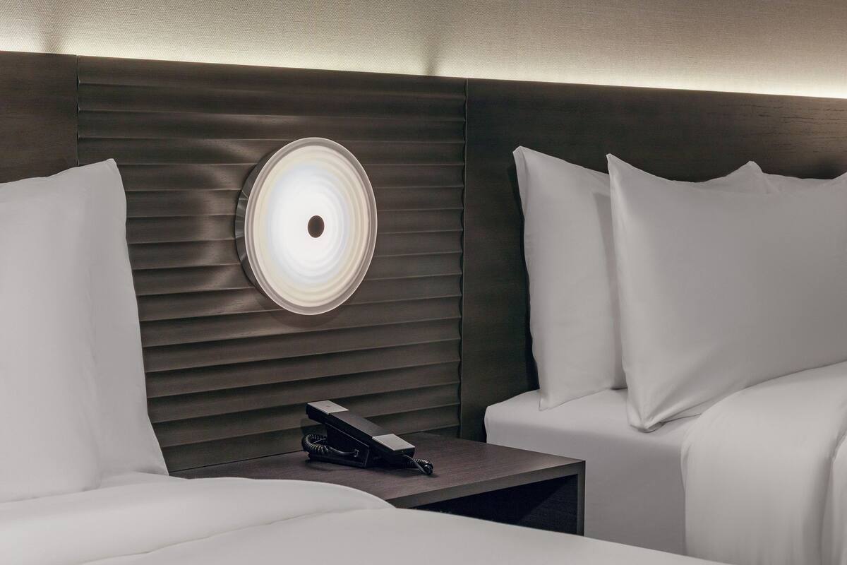 Habitación de hotel con diseño moderno, que incluye una cama con ropa blanca, una mesita de noche con teléfono, y una lámpara de pared.