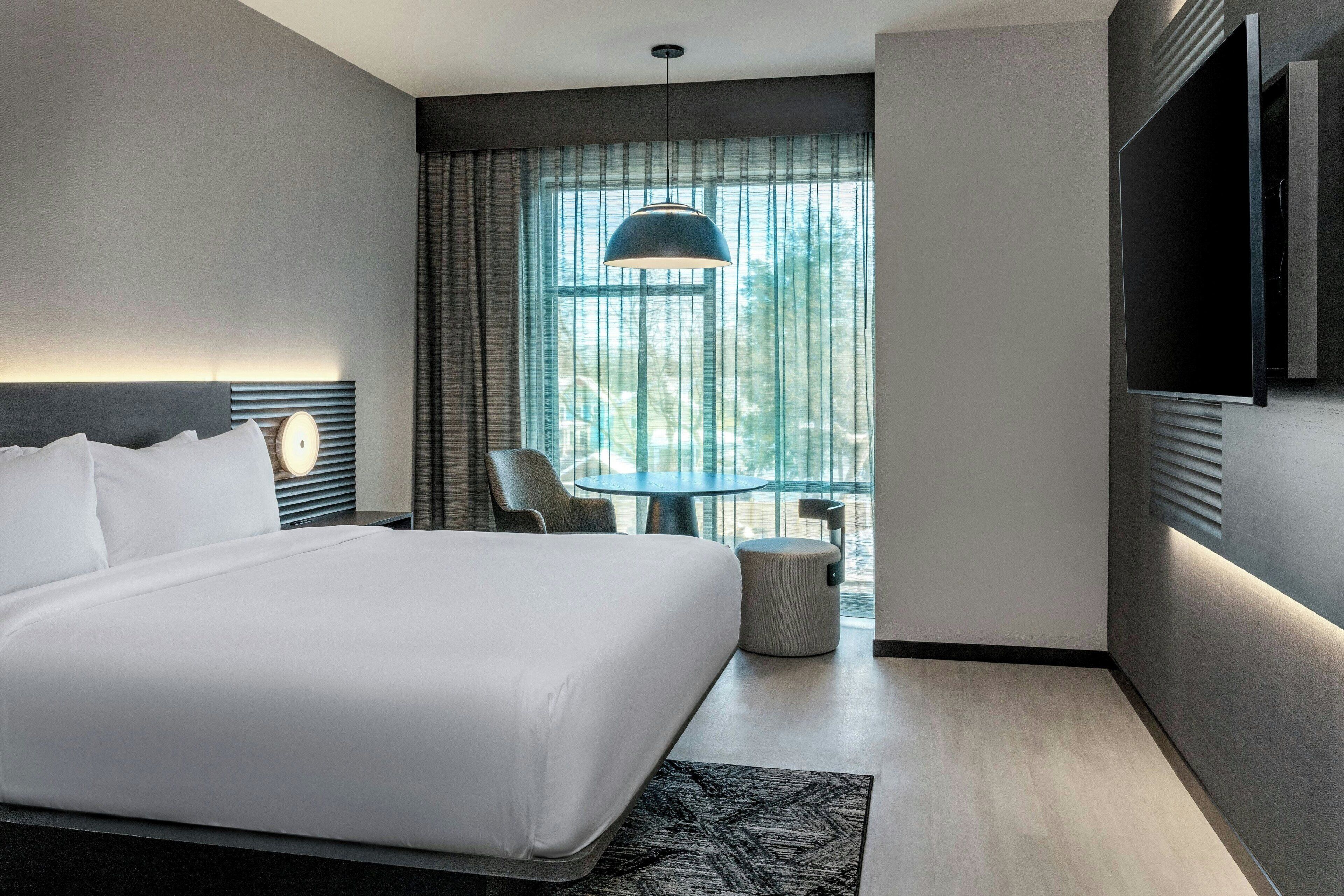 A modern hotel room with a large bed, a round table, a chair, a TV, and a large window with curtains.