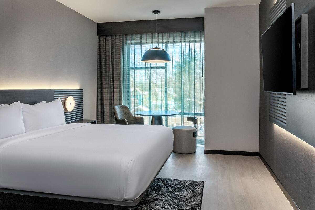 Una habitación de hotel moderna con una cama grande, una mesa redonda, una silla, un taburete pequeño, un televisor montado en la pared y una ventana con cortinas.