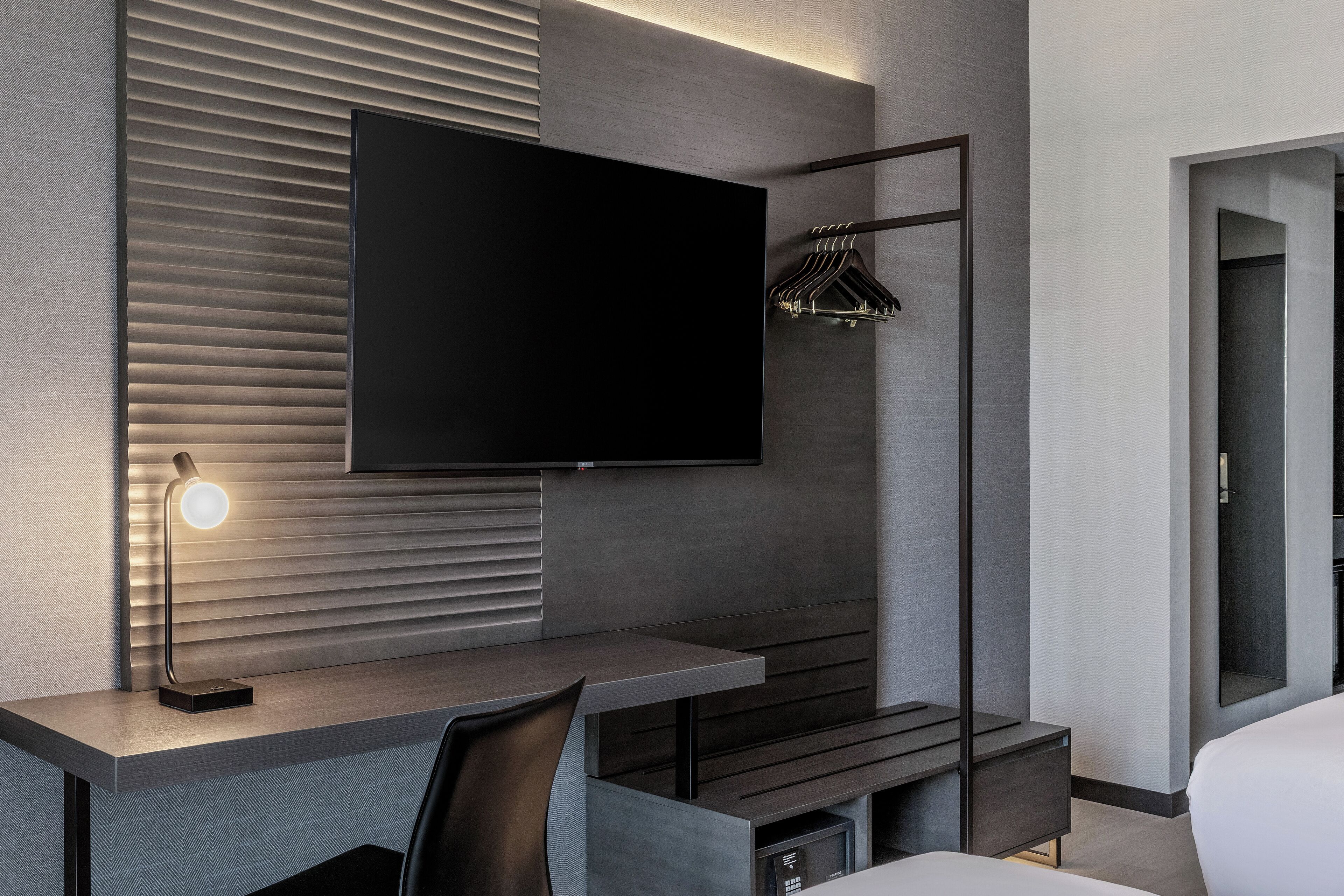 A modern hotel room with a flat-screen TV mounted on the wall, a desk with a lamp, and a clothing rack.