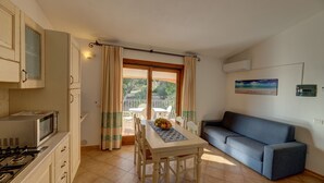 A compact kitchen with a microwave, stove, and cabinets. There's a dining table with chairs and a fruit bowl. A sofa is against the wall, and a window with curtains is visible. - One bedroom apartment (4 people) - Residence Giardini di Badus (Badesi)