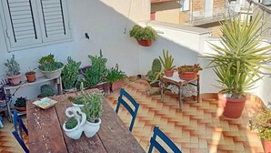 En udendørs terrasse med et træbord og blå stole, omgivet af krukker med planter.
