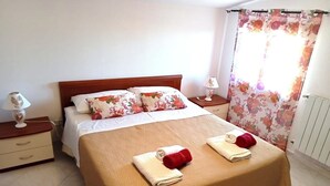 En pænt redt seng med blomsterpuder, et natbord med en lampe, og et vindue med blomstrede gardiner.