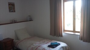 Een slaapkamer met een bed, een nachtkastje, een raam met gordijnen en een wandplank met flessen.