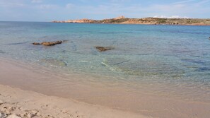 Una playa con aguas cristalinas y poco profundas, afloramientos rocosos y un paisaje montañoso distante.