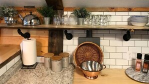 Une cuisine avec une étagère en bois où sont rangés divers ustensiles, un panier en osier et une passoire en cuivre posée sur le plan de travail.