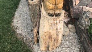 Deux sculptures d’ours en bois, posées sur des souches d’arbres, dans un espace jardin agrémenté d’un mur de pierre et d’un bâtiment en bois en arrière-plan.
