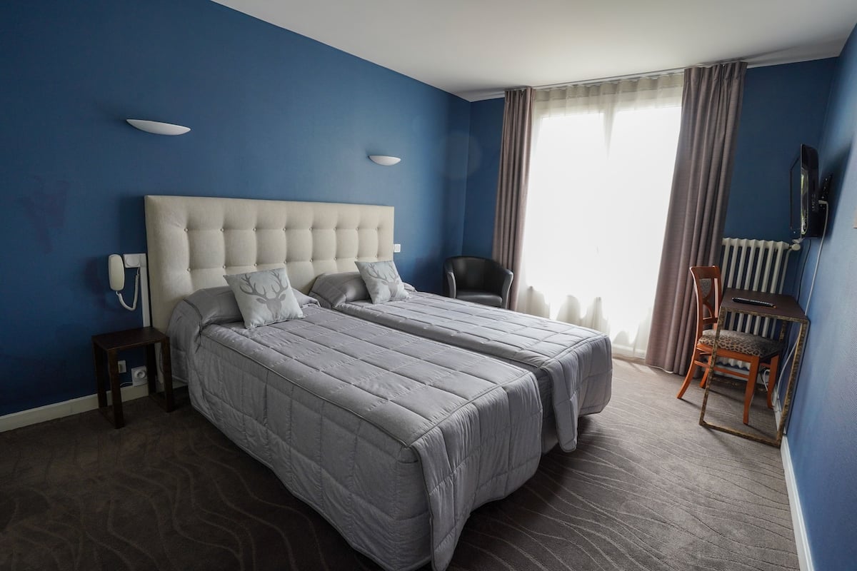 Ein Hotelzimmer mit zwei Betten, einem Sessel, einem Schreibtisch, einem Fernseher und einem Fenster mit Vorhängen.