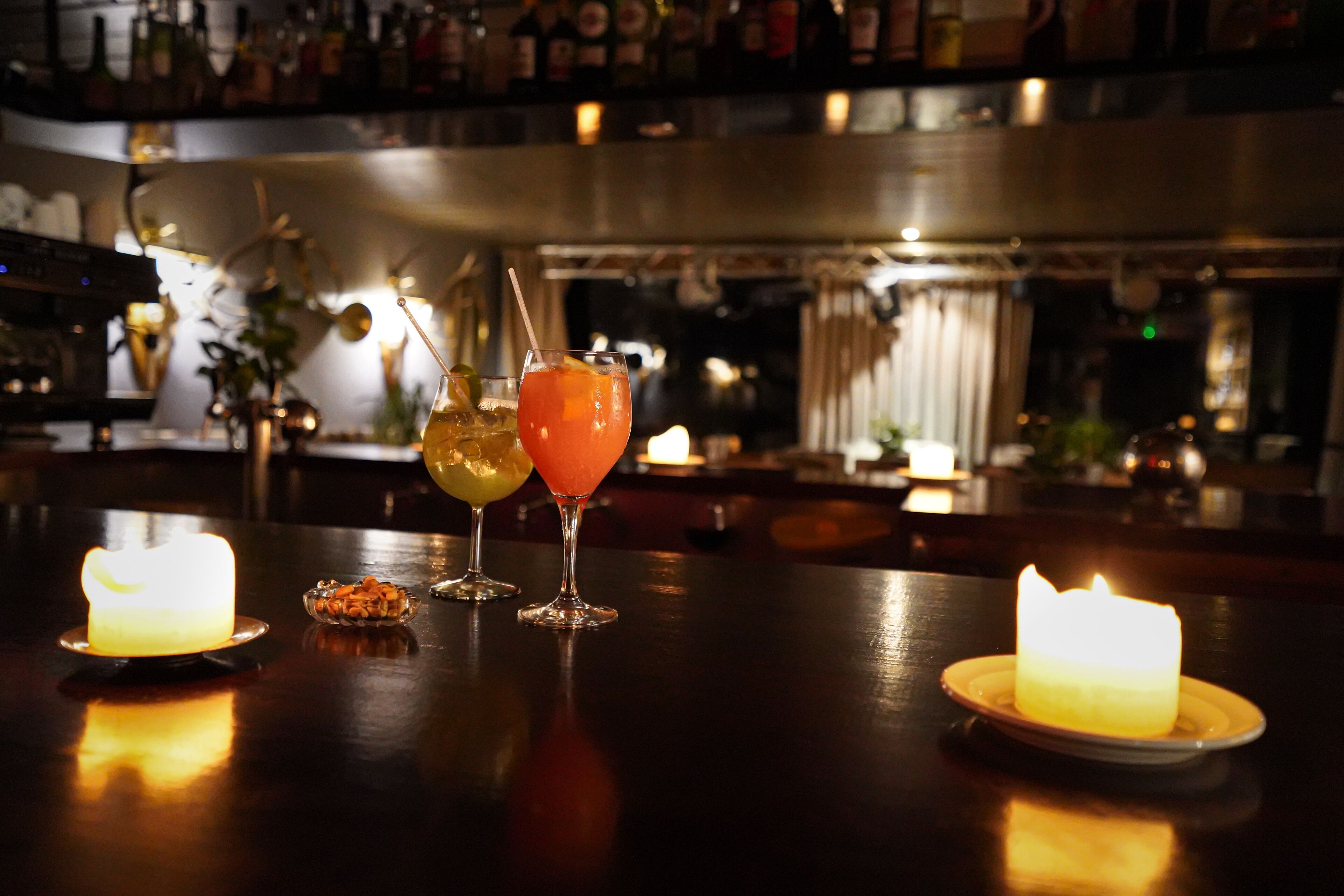 A bar area with three drinks on a table, a lit candle, and a bar stocked with bottles.