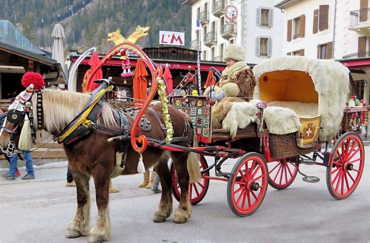Une calèche tirée par un cheval brun à la robe blanche sur les jambes, avec un siège bordé de fourrure et un cadre rouge, se déplace dans un paysage urbain, avec des bâtiments et une montagne en arrière-plan.