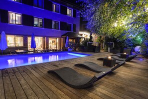 A nighttime pool area with lounge chairs, umbrellas, and a building with lit windows.