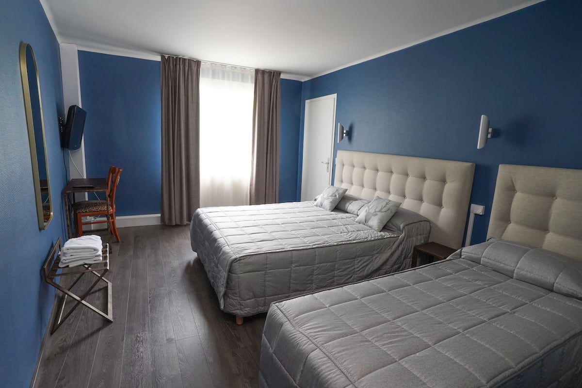 Ein Hotelzimmer mit zwei Betten, einem Fernseher, einem Sessel und einem Fenster mit Vorhängen.