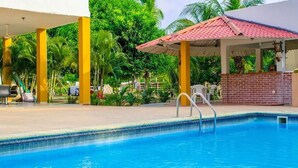 A swimming pool with a tiled deck, surrounded by palm trees and a covered area with a red roof. - Hotel San Basilio de Palenque (Mahates)