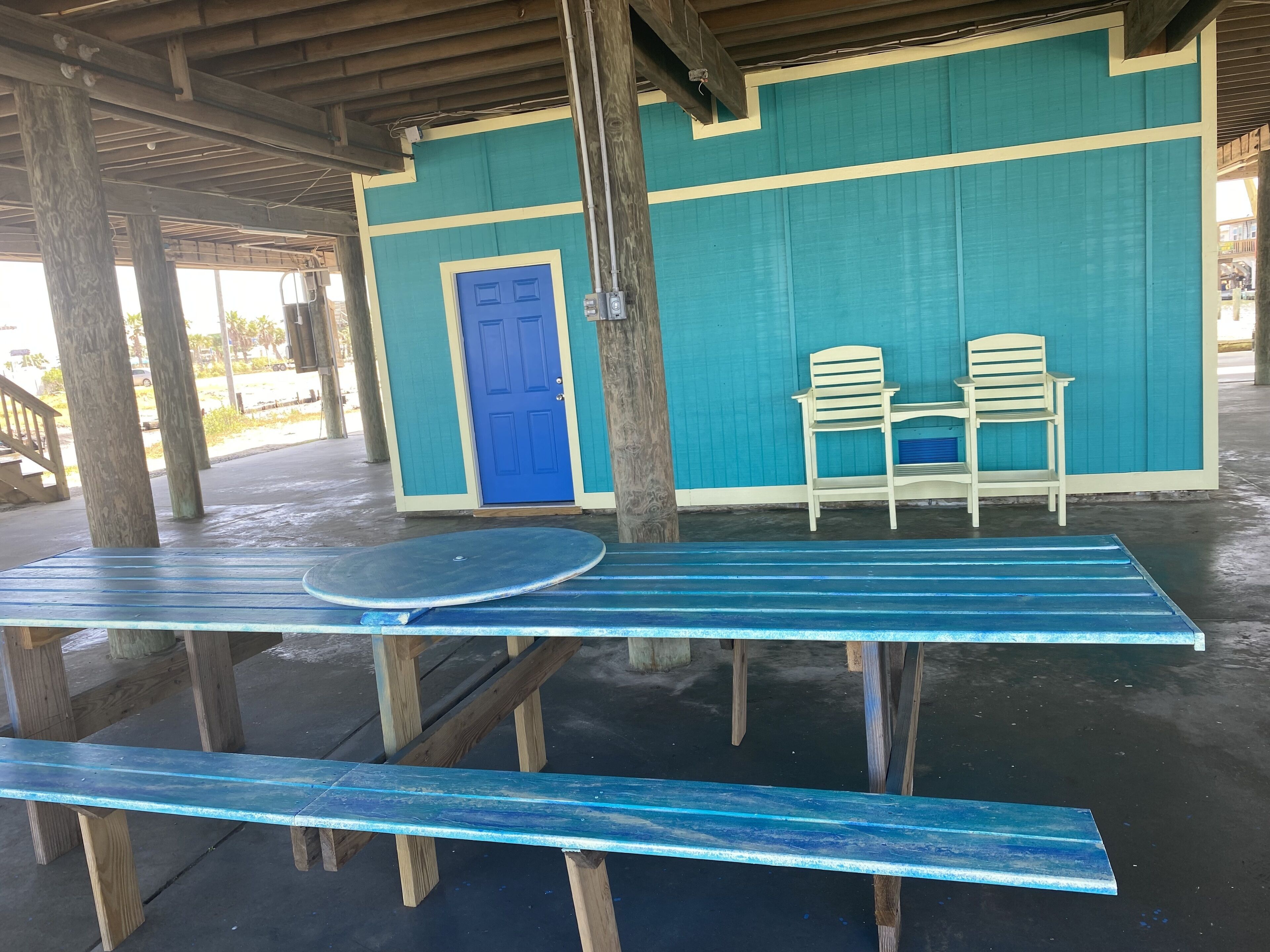 A covered outdoor seating area with a blue table and benches, a blue door, and a teal wall.