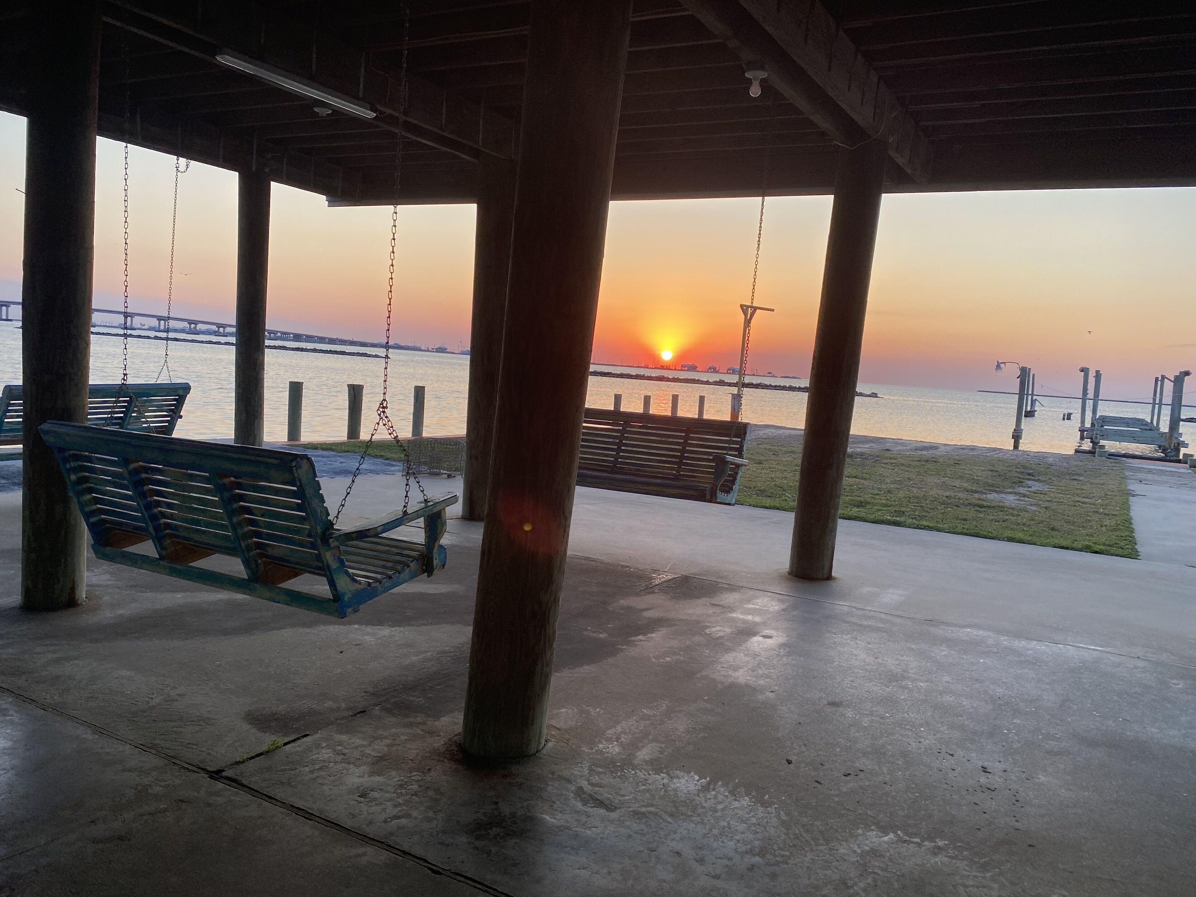 Un espace extérieur abrité, doté d’une balançoire et d’un banc, d’où l’on peut admirer un coucher de soleil sur une étendue d’eau, avec un pont visible au loin.