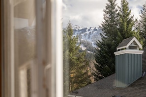 A view of a mountain range with snow-capped peaks, dense evergreen trees, and a small green-roofed structure with a white trim. - 2BR | Granite Court | Village Location | King Bed | Private Balcony | Parking | Fireplace (Whistler)