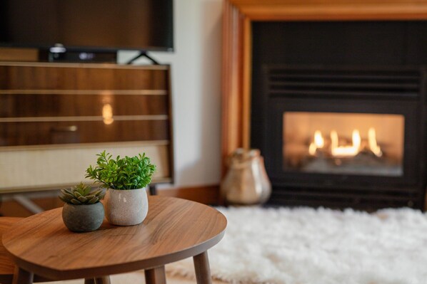 A cozy living room with a wooden table, two potted plants, a fireplace, and a television. - 2BR | Granite Court | Village Location | King Bed | Private Balcony | Parking | Fireplace (Whistler)