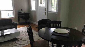 Un coin repas avec une table ronde, des chaises et une assiette blanche. On aperçoit, à l’arrière-plan, un canapé gris et un bureau avec une chaise.