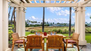 A covered outdoor dining area with a wooden table and chairs, a floral rug, and a view of a green landscape with palm trees.