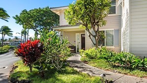 A two-story residential building with a front porch, a driveway, and a variety of plants and trees.