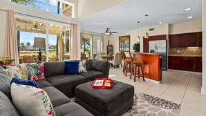 A spacious living room with a large gray sectional sofa, a dining area with a bar, and a kitchen with dark wooden cabinets.