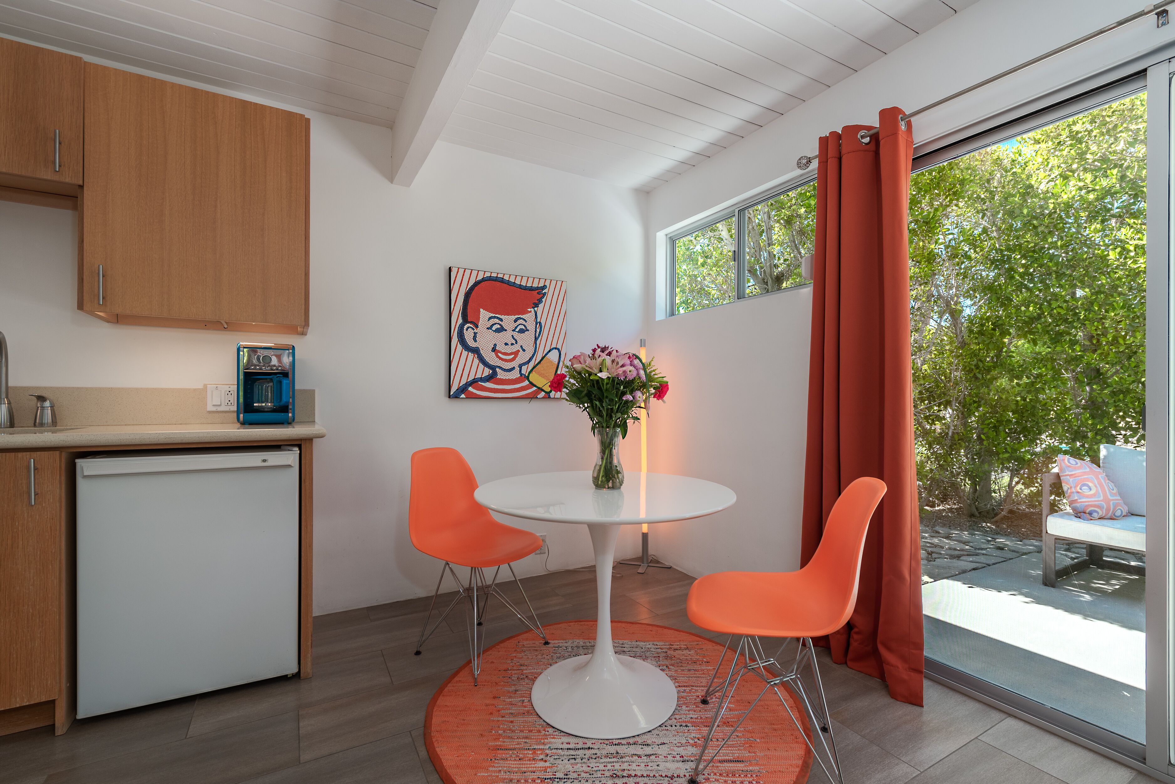 A compact kitchen with a white refrigerator, two orange chairs, a white table, and a vase with flowers.