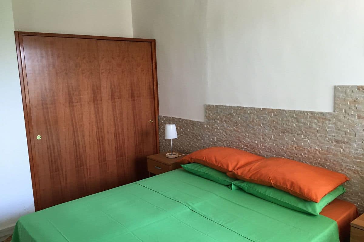Une chambre avec une armoire en bois, une table de chevet avec une lampe et un lit avec une literie aux couleurs verte et orange.