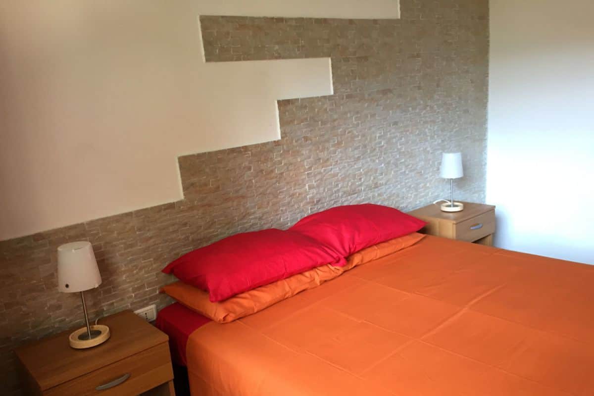 Une chambre à coucher avec une tête de lit ornée d’un motif en briques, un lit avec une literie orange, deux tables de chevet avec des lampes et une petite table de nuit avec une lampe.