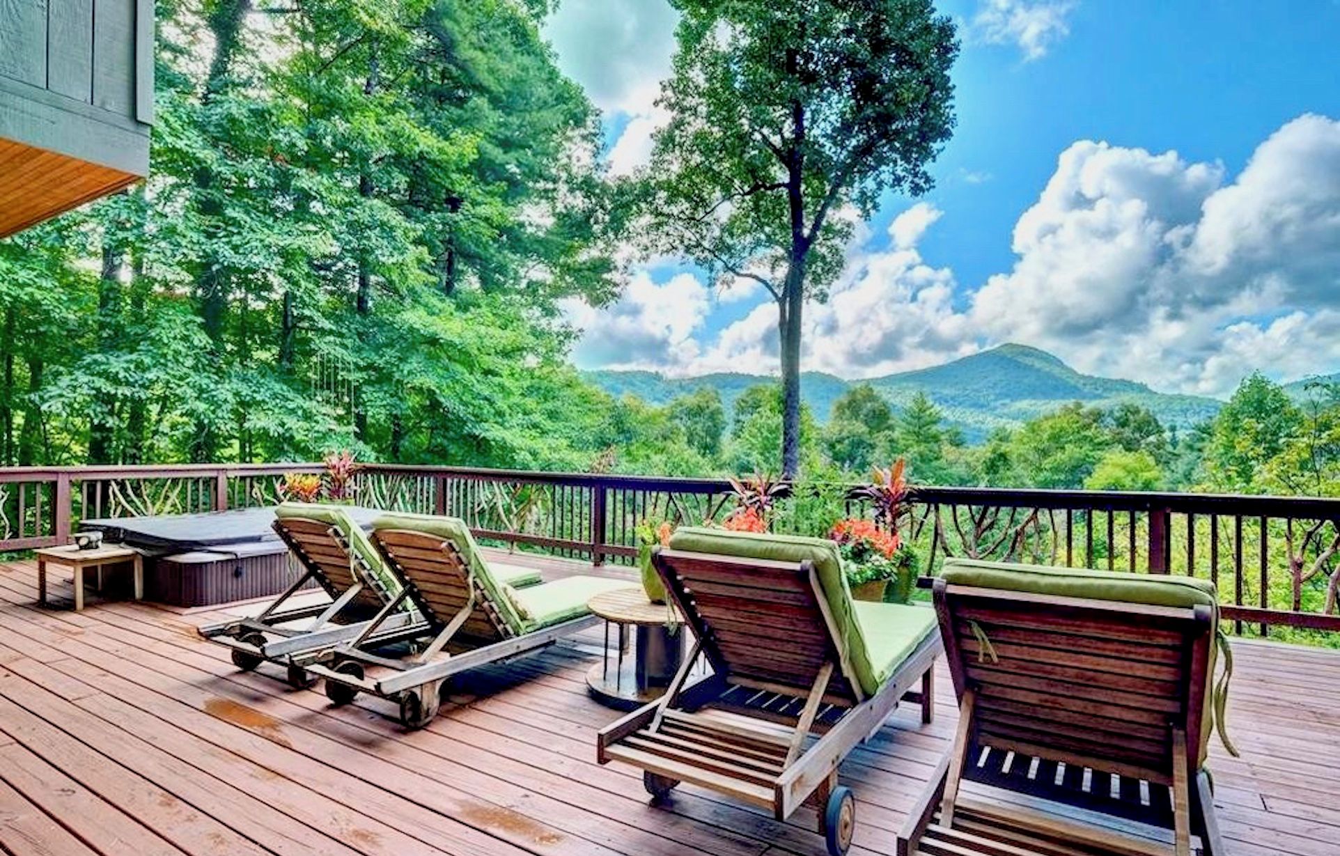 A wooden deck with lounge chairs and a small table, overlooking a lush green landscape with trees and a mountain in the distance.