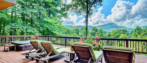 A wooden deck with lounge chairs and a small table, overlooking a lush green landscape with trees and a mountain in the distance.