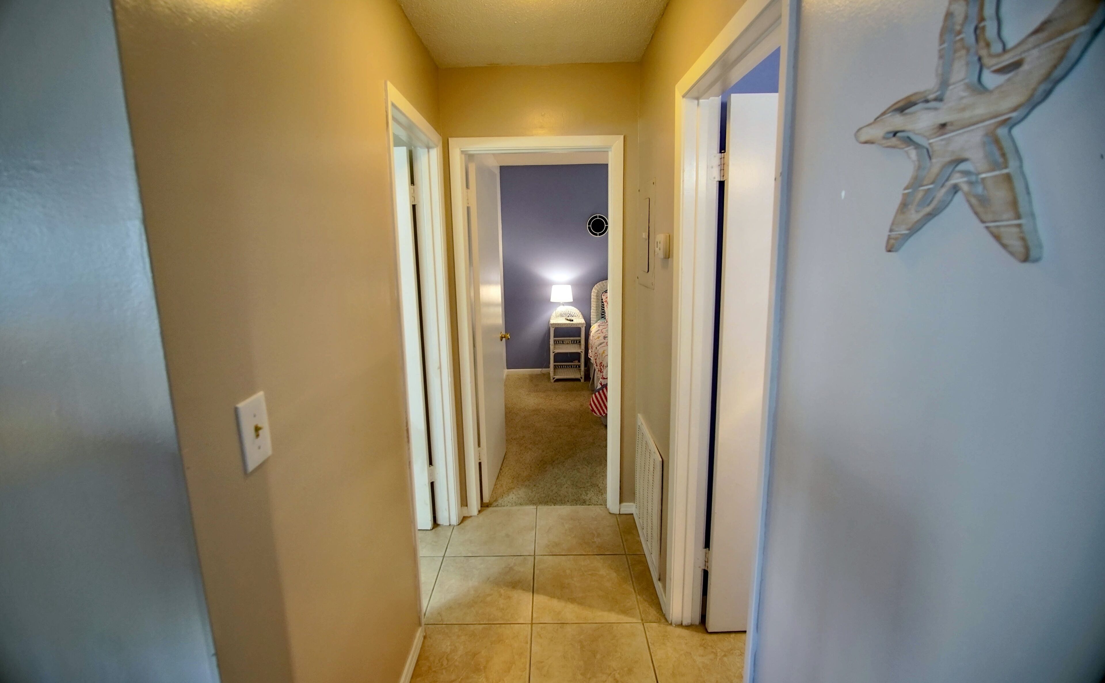 A narrow hallway with beige walls and a tiled floor leading to a bedroom.