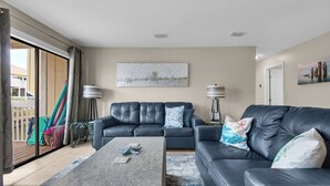Ein Wohnzimmer mit grauem Sofa, einem Couchtisch und zwei Stehlampen. Durch die Glastür ist ein Balkon zu sehen.
