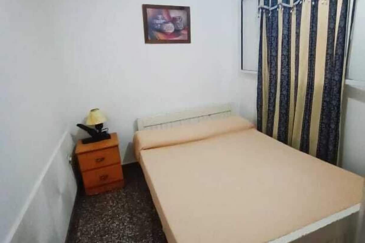 A small, single-bed room with a wooden bed frame, a small wooden nightstand with a lamp, a framed picture on the wall, and a striped curtain.